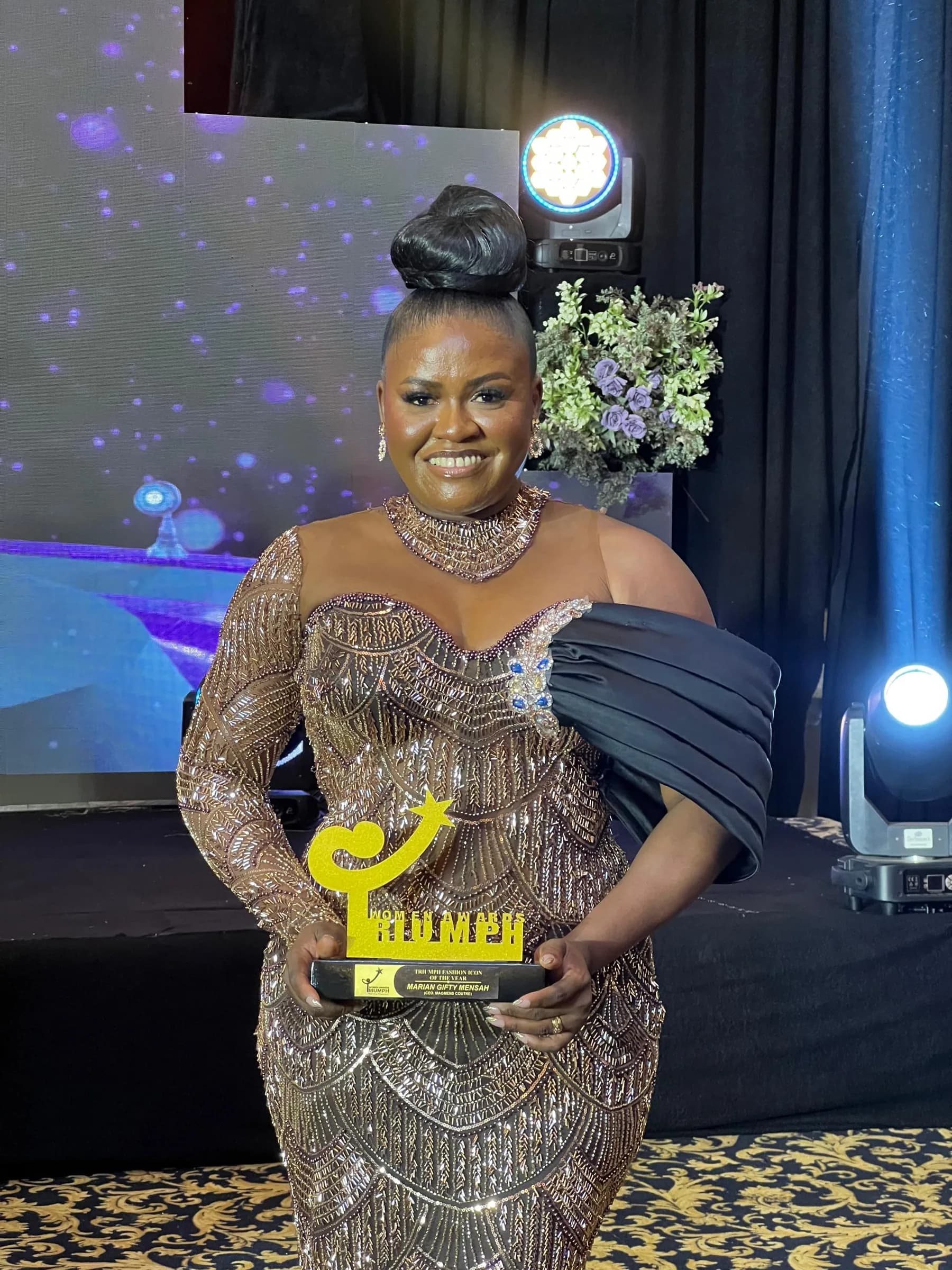 Mrs. Marian Gifty Mensah-Dossah, founder and CEO of Magmens Couture, accepting an award in 2026, holding a gold-and-black award plaque with her name on the base.
