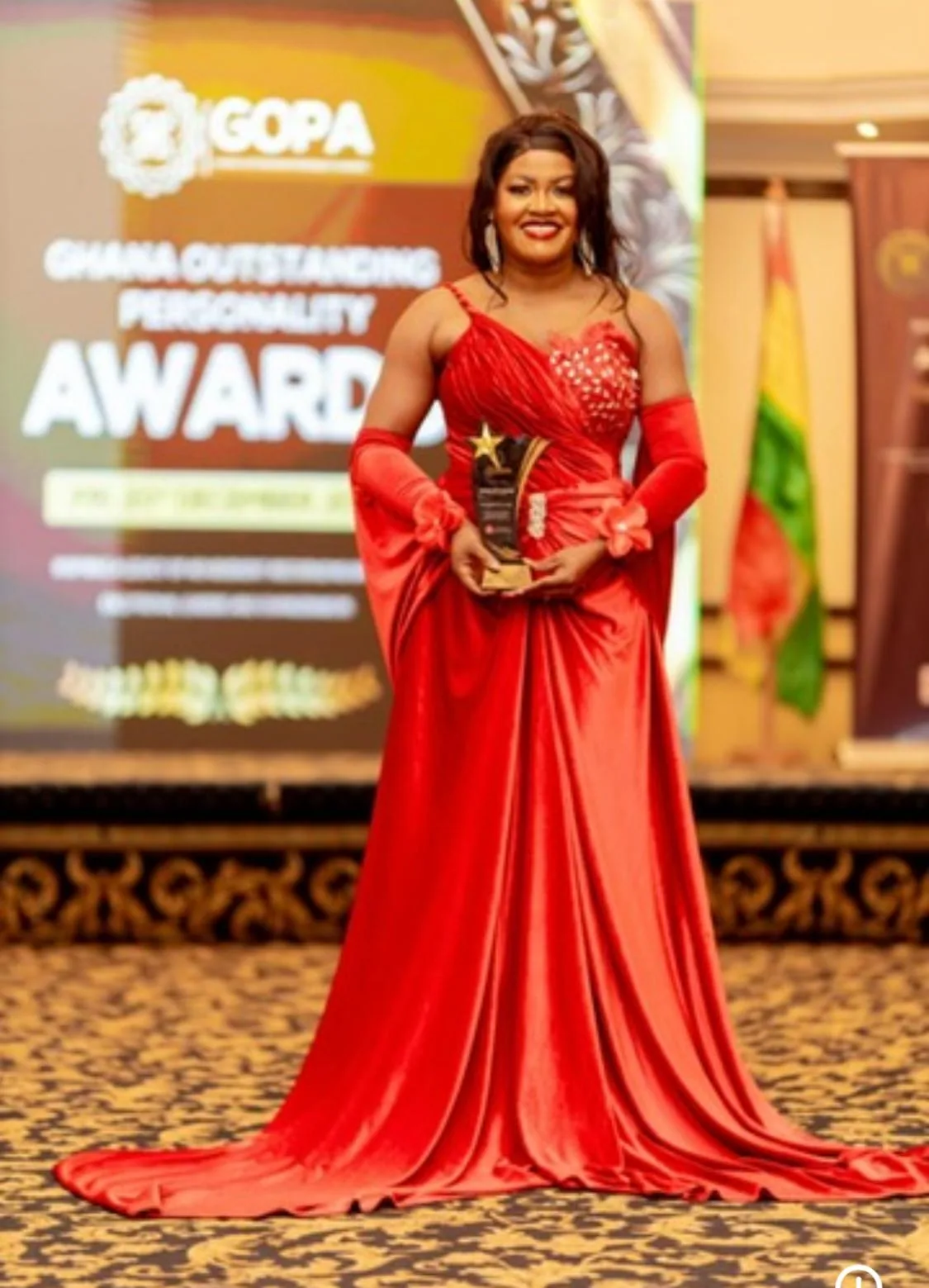 Mrs. Marian Gifty Mensah-Dossah in a red gown accepting an award at a stage ceremony, with a Ghanaian flag visible behind her.