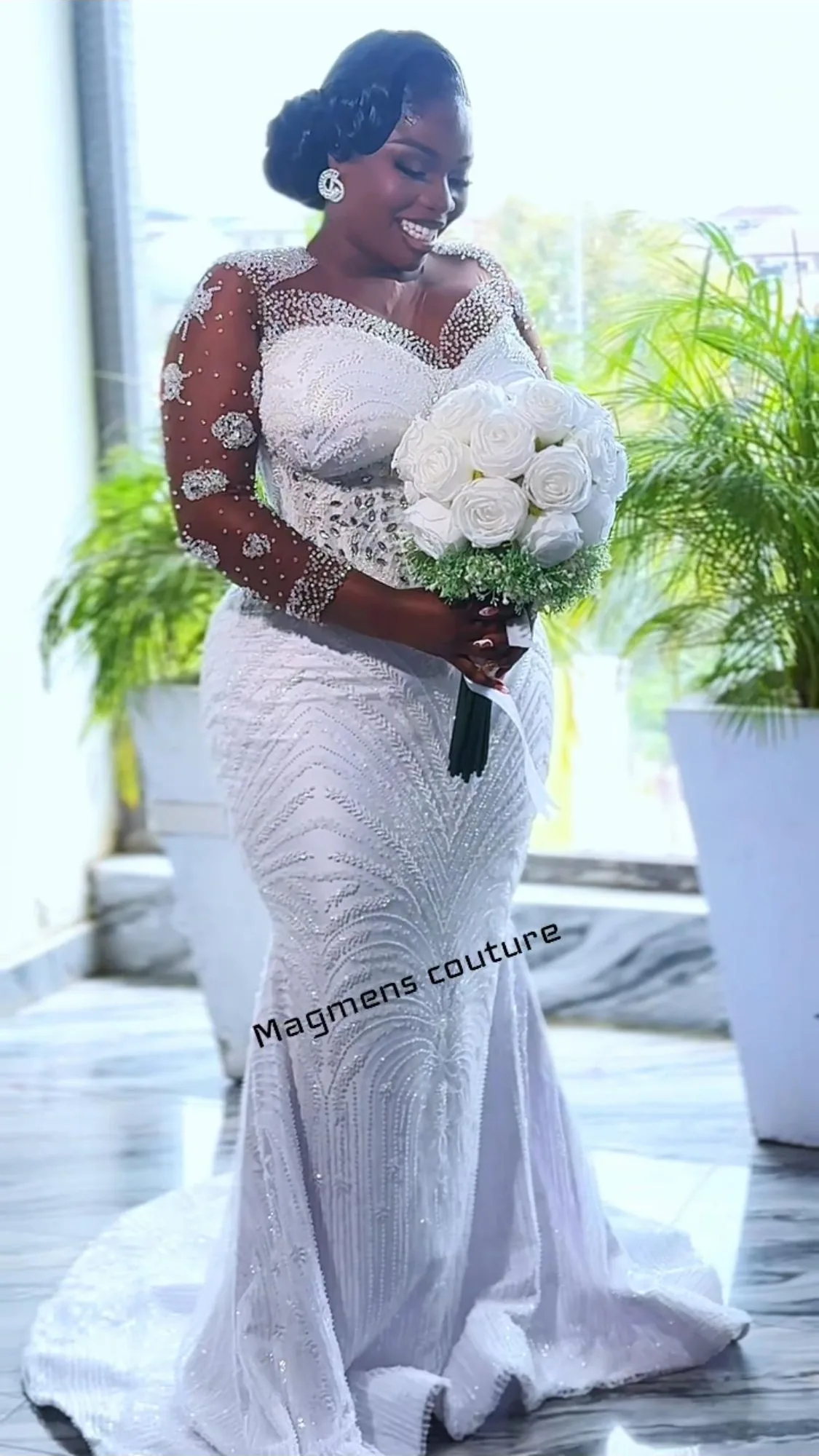 Bride in a long-sleeved beaded Magmens Couture mermaid gown with an illusion neckline and cathedral train, holding a bouquet of white roses beside a plant-filled window.