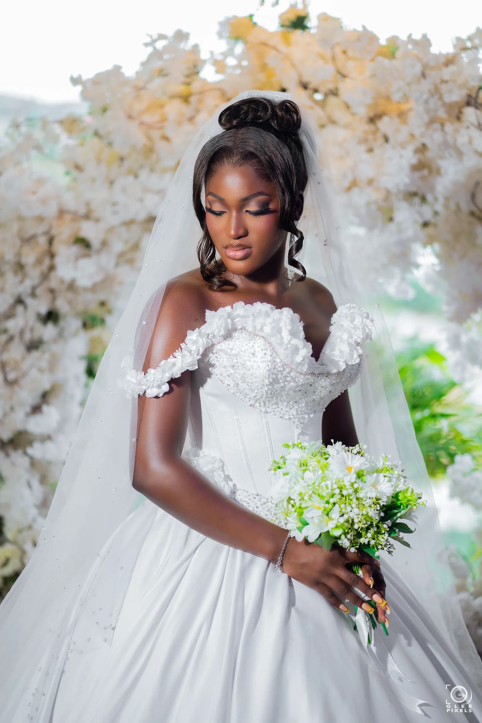 Bride wearing a custom ivory satin Magmens Couture ball gown with a sculpted off-shoulder floral lace bodice, photographed before an installation of cream blossoms.