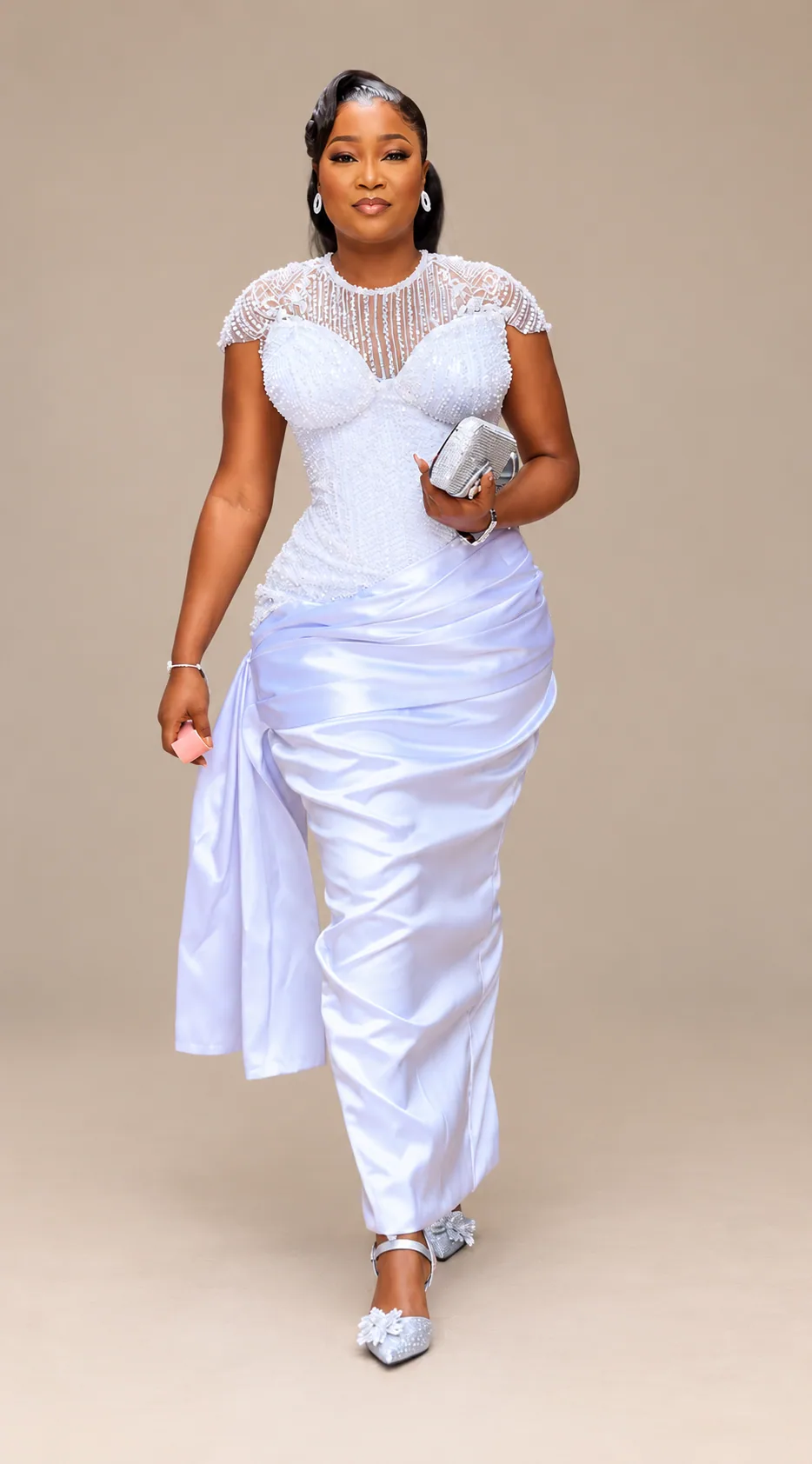 Client in a white hand-beaded corset with a sheer yoke and cap sleeves, worn with a draped white satin skirt and a silver clutch, photographed in a pale interior.
