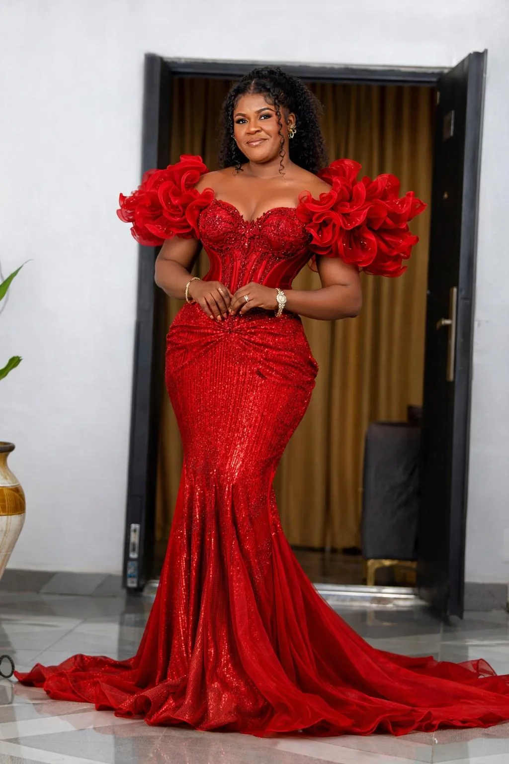 Mrs. Marian Gifty Mensah-Dossah, founder and CEO of Magmens Couture, in a custom red sequin mermaid gown with sculpted ruffle off-shoulder sleeves, a corseted bodice and a sweeping train, photographed in a doorway.