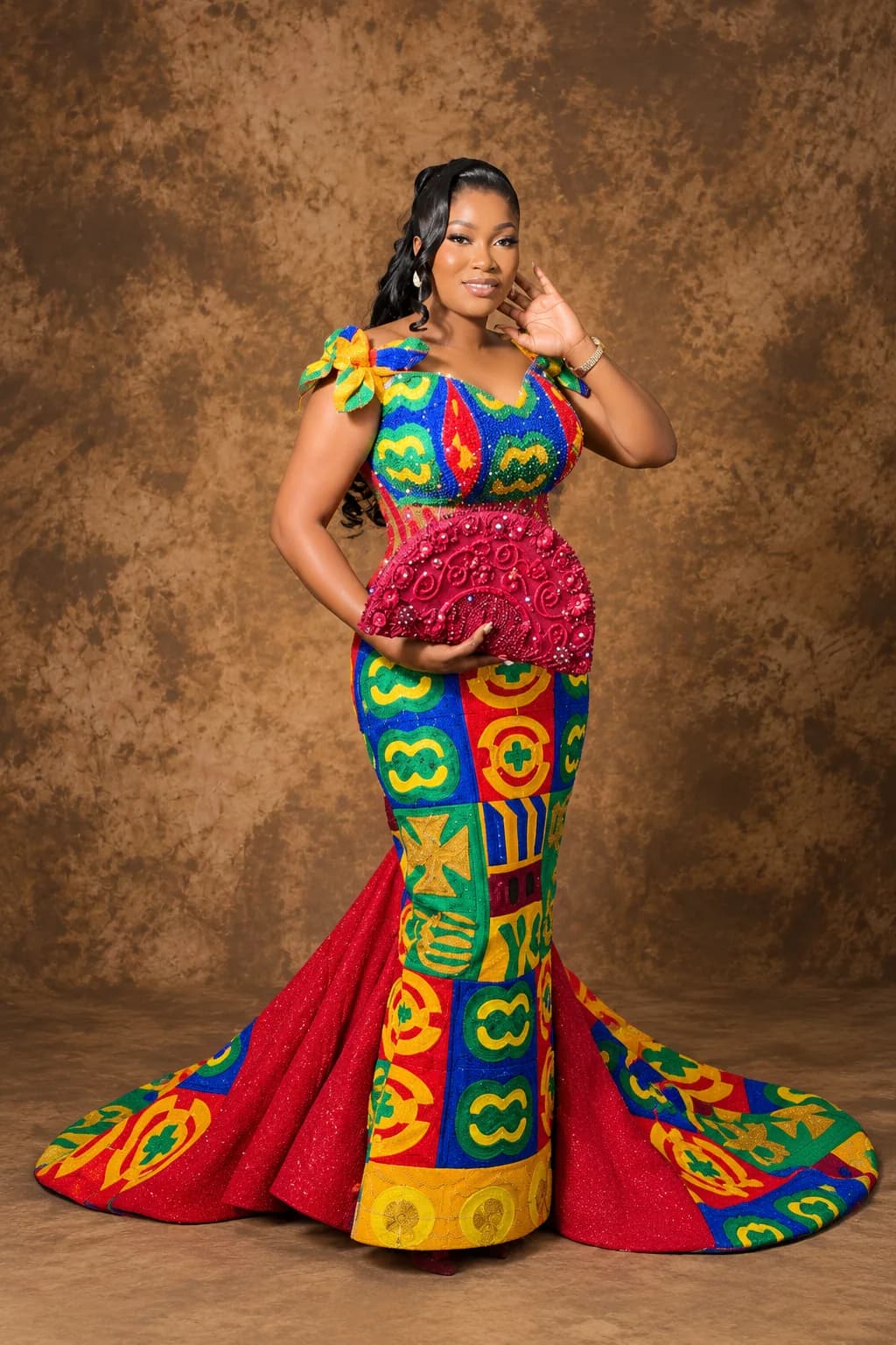 Client in a beaded multi-colour Kente gown with a coral fishtail train, holding a deep red beaded clutch, photographed against a bronze studio backdrop.