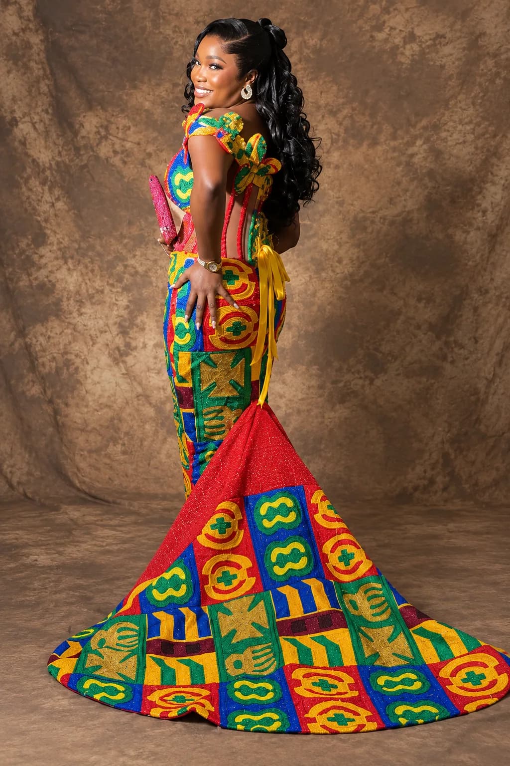 Client seen from behind in a hand-beaded Kente corset and matching Kente fishtail skirt tied with a red sash, the train fanned across the studio floor.